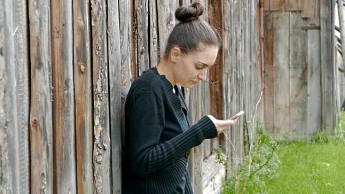 年轻女孩靠在一个旧谷仓上，用她的智能手机