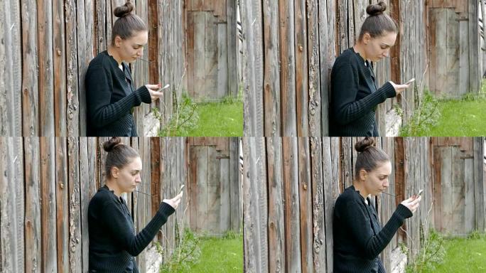 年轻女孩靠在一个旧谷仓上，用她的智能手机