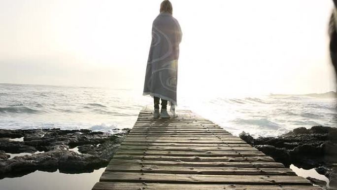 一名妇女裹着毯子沿着海边的木板路行走