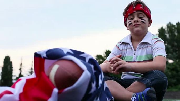 男孩坐在美国国旗的美式橄榄球旁边