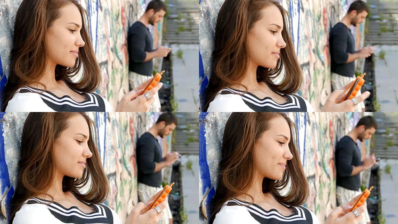 年轻的成年男女靠在涂鸦墙上并使用智能手机