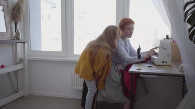 美丽的红发妈妈缝制衣服，坐在窗户附近的桌子旁，向女儿展示如何制作正确的接缝。