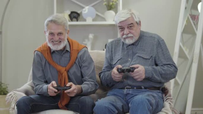 两个快乐的白人老人玩电子游戏，给了五个，微笑着。积极开朗的退休人员在室内玩得开心。