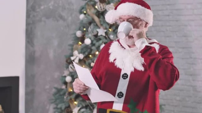 穿着圣诞老人服装的白人男子微笑着喝咖啡，读信。戴眼镜的圣诞老人站在圣诞树的背景下微笑。电影院4k P