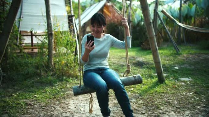 迷人的女人坐在秋千上，在后院使用智能手机