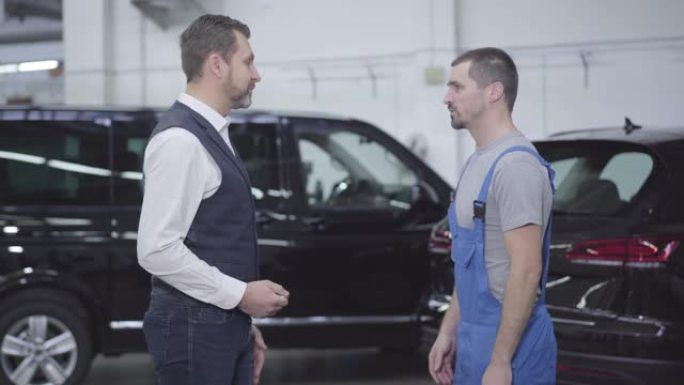 成年高加索商人站在汽车修理厂与工人交谈的侧视图。英俊的成功男人向汽车修理工咨询他的汽车。维修服务。