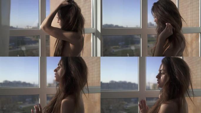 年轻裸体美丽性感黑发长发站在房子窗户附近。女性抚摸她浓密的头发。