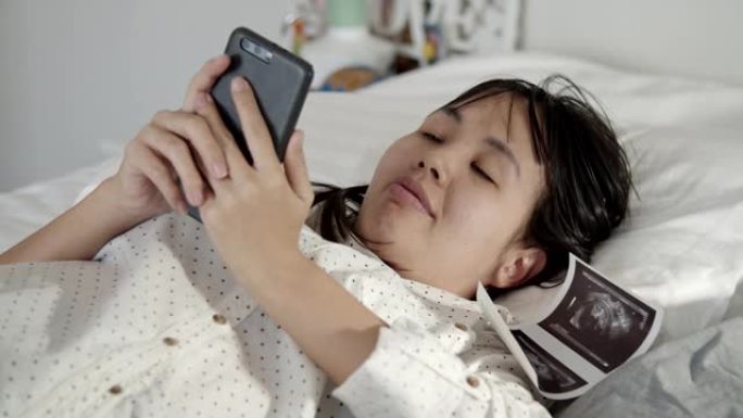 幸福孕妇躺在床上一边看手机上的3D超声波视频