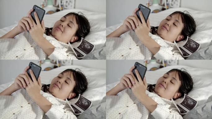 幸福孕妇躺在床上一边看手机上的3D超声波视频