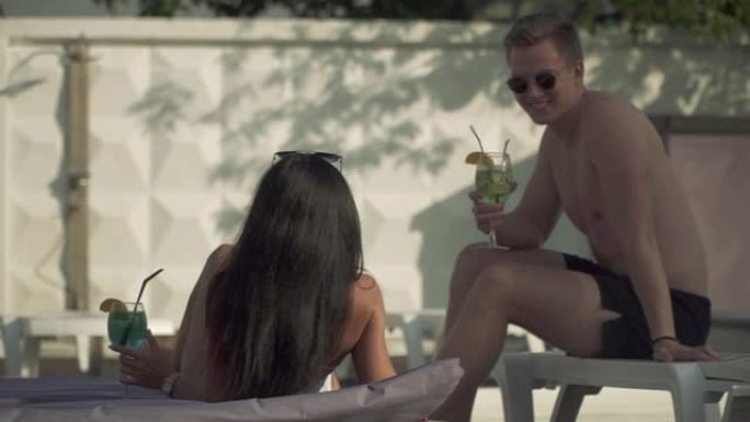 一个穿着泳裤和太阳镜的年轻人与两个穿着泳衣的漂亮女友交流，这些女友坐在室外游泳池附近的日光浴床上。男