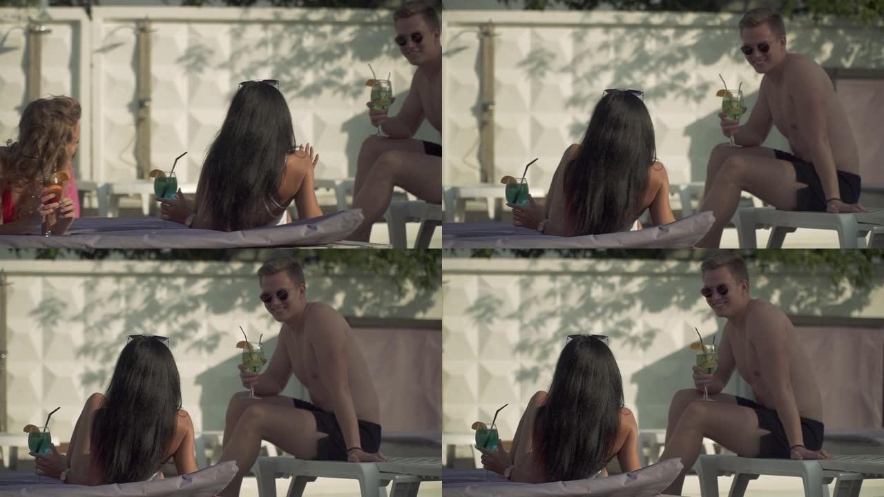 一个穿着泳裤和太阳镜的年轻人与两个穿着泳衣的漂亮女友交流，这些女友坐在室外游泳池附近的日光浴床上。男