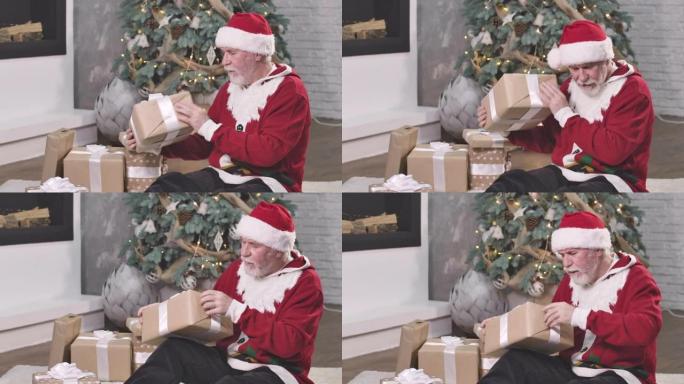 白胡子和胡须的白人老人拿着礼品盒并摇晃它。戴着红帽子和圣诞毛衣的圣诞老人坐在新年树下的地板上，带着礼