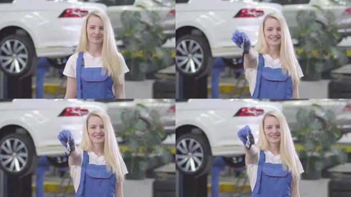 开朗的白人妇女站在汽车的背景下，向相机展示车钥匙。金发女汽车修理工对她的工作很满意。生活方式，职业，