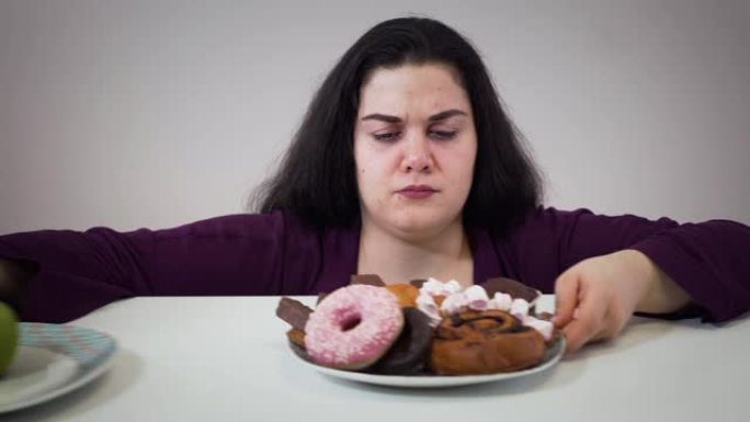 胖胖的白人妇女将苹果放在盘子旁，吃不健康的糖果。有超重问题的微笑丰满女孩的肖像。肥胖，不健康的生活方