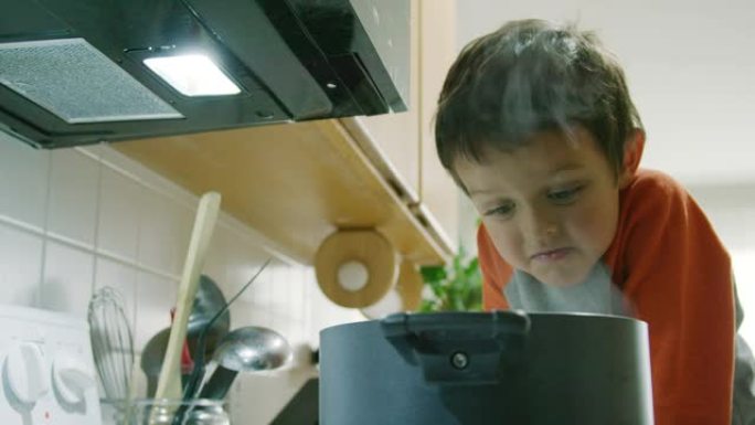 一个五岁的高加索男孩表现得很傻，对着一个热气腾腾的锅做鬼脸，锅坐在厨房的炉灶上