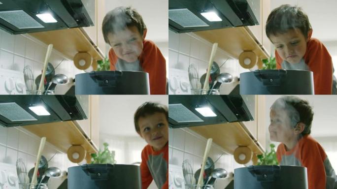 一个五岁的高加索男孩表现得很傻，对着一个热气腾腾的锅做鬼脸，锅坐在厨房的炉灶上