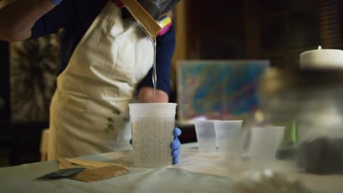 一名戴着安全手套和呼吸器的妇女在室内艺术工作室用木制工艺棒搅拌透明的环氧/树脂