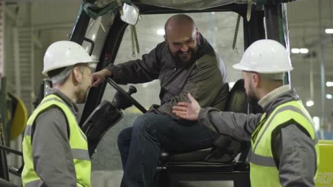 两名戴着头盔和背心的白人男性工人与坐在牵引车上的大胡子同事交谈。工厂员工在室内工作。制造、装载机、生