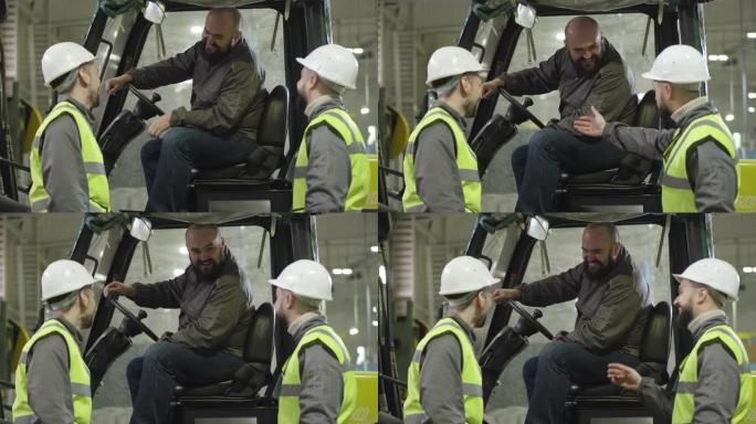 两名戴着头盔和背心的白人男性工人与坐在牵引车上的大胡子同事交谈。工厂员工在室内工作。制造、装载机、生