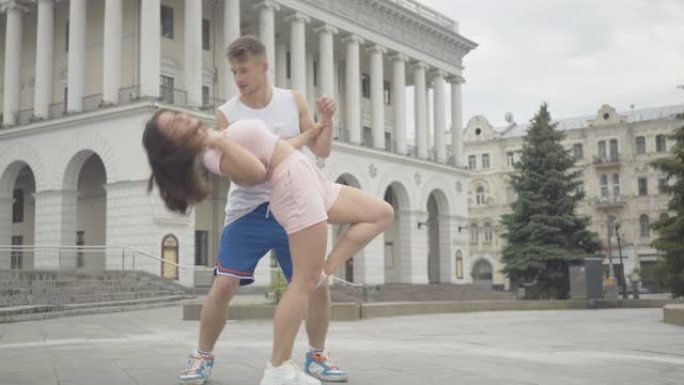 相机接近自信的男人旋转和弯腰年轻美丽的女人。在市中心广场表演的专业舞者肖像。高加索夫妇在户外跳舞。