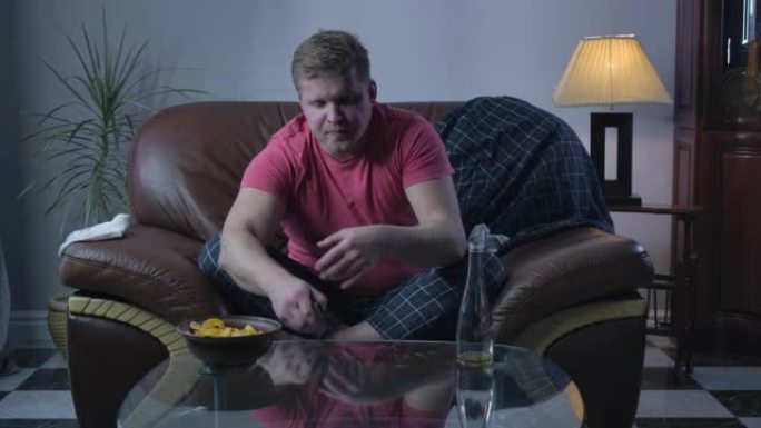 沮丧的白人男子脱下袜子并用遥控器切换频道的肖像。单身懒汉拿着薯条和啤酒看电视。生活方式，休息，喝酒。
