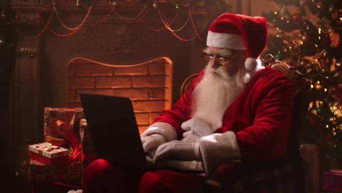 圣诞老人的魔术室坐在家里的光束中，在圣诞节前夕，壁炉和圣诞树的背景下，坐在互联网上的笔记本电脑上打字