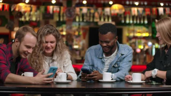 坐在咖啡馆里的四个朋友笑着看着手机屏幕，交换旅行中的照片。非裔美国女友聊天喝咖啡。