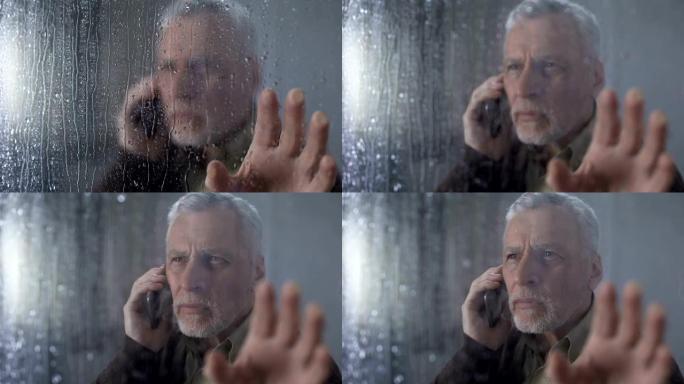 担心的老人透过下雨的窗户，打电话，等待