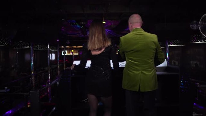 女性高加索DJ混合音乐的后视图，老年男性访客在控制器旁边跳舞。穿着黑色连衣裙的年轻女子和穿着绿色夹克
