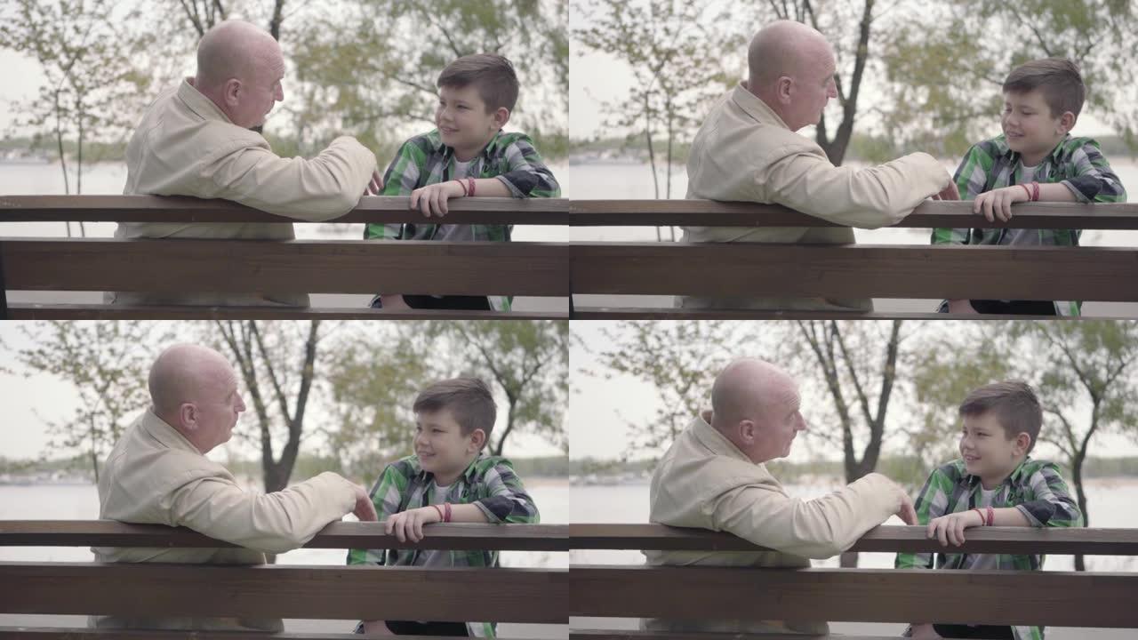 肖像祖父和孙子坐在河边的公园里的长凳上，老人给男孩讲有趣的故事