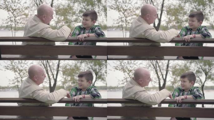 肖像祖父和孙子坐在河边的公园里的长凳上，老人给男孩讲有趣的故事