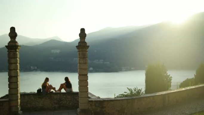 两个女孩坐在石墙上，被柱子框住，俯瞰湖泊和山脉