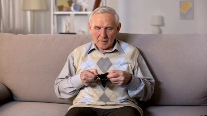 悲伤的老人坐在沙发上，在镜头前露出空钱包，贫穷