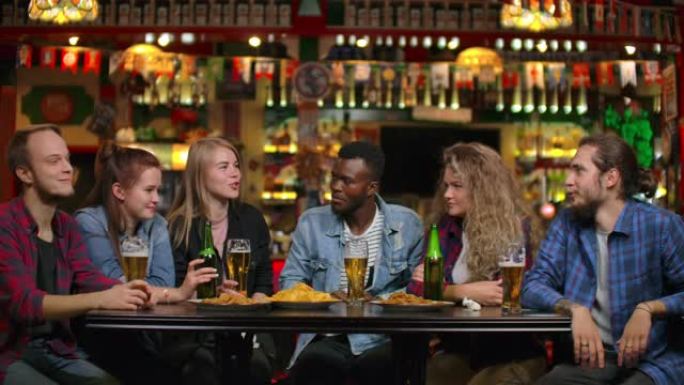 一群多种族的年轻男女在酒吧喝啤酒，吃薯条，兴高采烈地辩论大学。嘲笑一个笑话