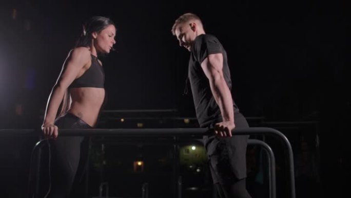 在慢动作中，一个男人和一个女人在城市公园的黑暗中一起在双杠上进行俯卧撑。夜间训练
