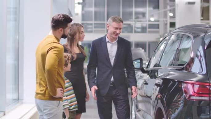 自信的男性汽车经销商在汽车陈列室向高加索家庭宣传新车，并与年轻人握手。丈夫、妻子和儿子购买新车。电影