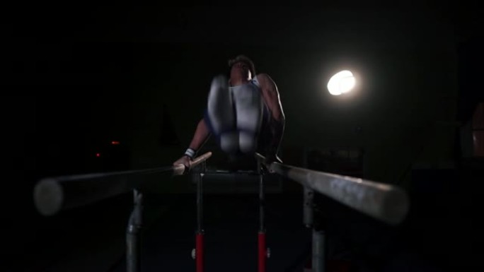 男体操运动员杂技演员在黑暗的房间中以慢动作在双杠上倒立，分享翻筋斗并降落在地板上。奥运会前的训练。职