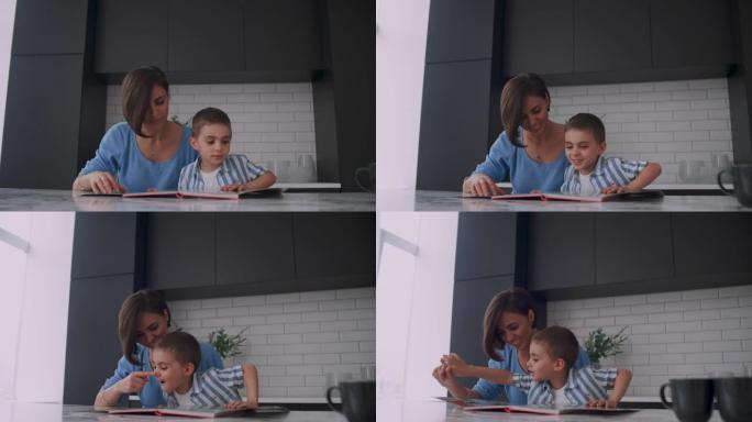 年轻美丽的母亲和儿子坐在明亮的厨房里的桌子旁看书，看着照片，用手指戳进书本，翻阅书页。