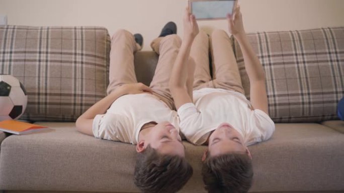 两个开朗的高加索双胞胎兄弟躺在沙发上，使用平板电脑。兄弟姐妹一起在家休息。儿童使用社交网络，游戏成瘾
