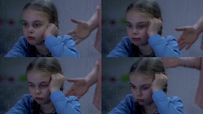 女士对坐在下雨的窗户附近的哭泣的小女孩大喊大叫，羞辱孩子