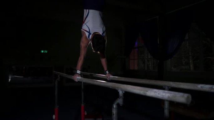 男体操运动员杂技演员在黑暗的房间中以慢动作在双杠上倒立，分享翻筋斗并降落在地板上。奥运会前的训练