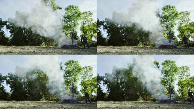 四十多岁的高加索人将树叶，木棍和树枝和灰烬耙到户外的一个大燃烧燃烧堆上，背景是大树