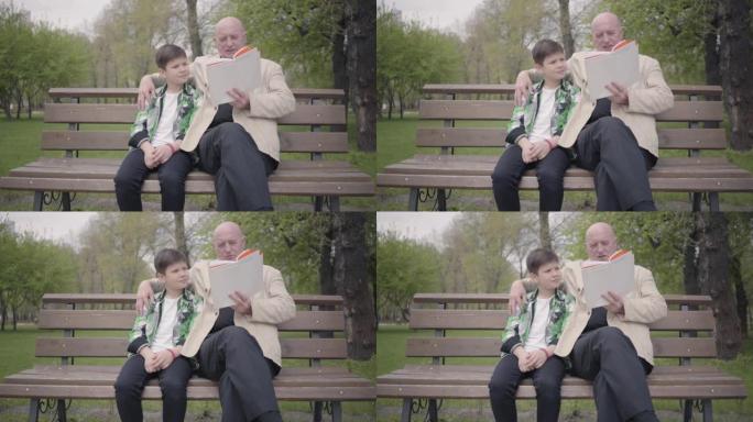 肖像可爱的爷爷和可爱的孙子坐在公园的长凳上，老人为男孩看书。