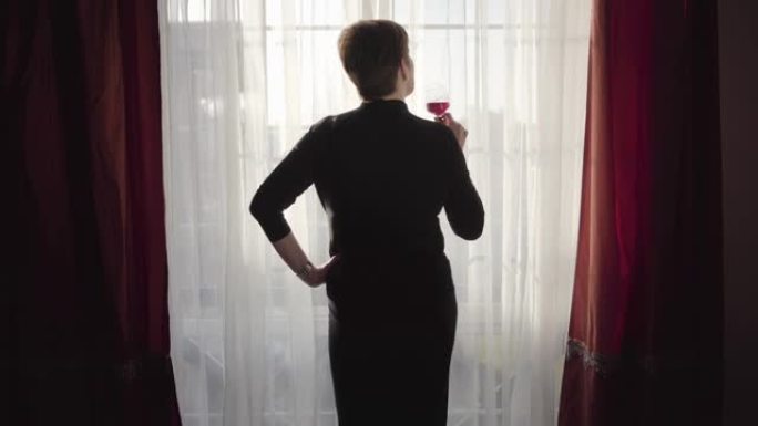 穿着优雅黑色连衣裙的黑发高加索女人站在大窗户前，一只手放在臀部，喝着红酒。自信的女商人在晚上辛苦工作