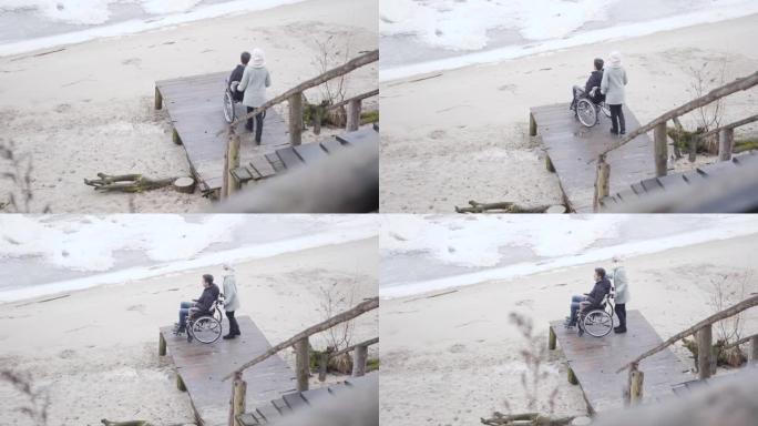 年轻强壮的母亲带着残疾的儿子沿着码头滚动轮椅，与残疾的年轻人交谈。高加索人无效的嫩花秋日与病人在户外