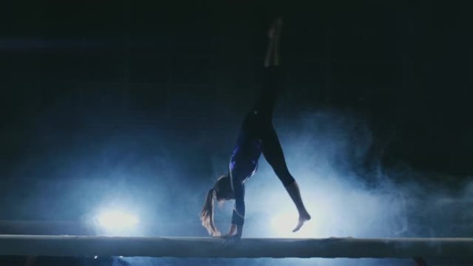 女孩在背光和运动体操服装中的慢动作上表演技巧。烟雾和蓝色。在平衡木上跳跃和旋转