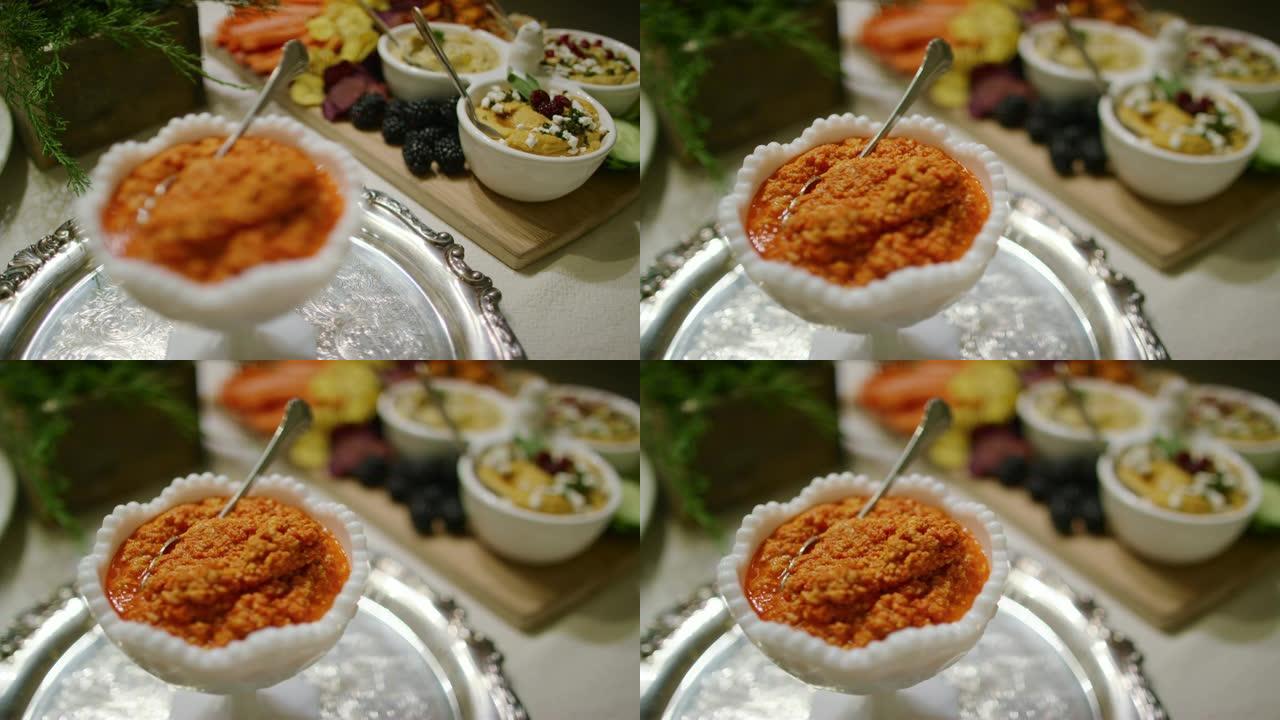 在室内庆祝活动/聚会的桌子上，一碗橙色的Romesco蘸酱放在银色的盘子上，上面放着各种蘸酱，开胃菜