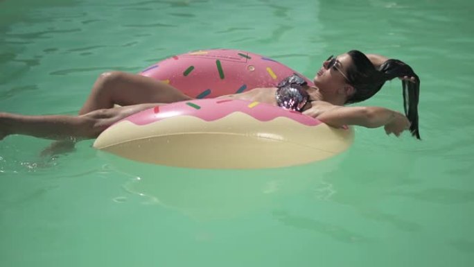 戴着墨镜的漂亮女人躺在粉红色的橡胶圈上享受着阳光在游泳池里游泳。穿着比基尼的寂寞女人的休闲。