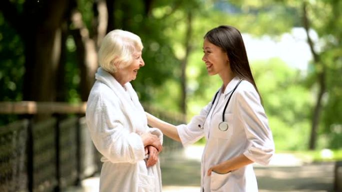 微笑的护士在sanatoria公园与穿着浴袍的老年妇女交谈，放松