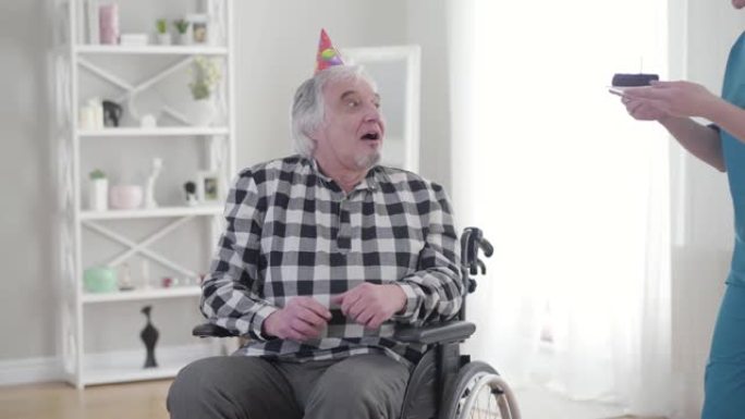 坐在轮椅上的快乐白人老人从无效的温柔那里收到生日蛋糕的肖像。快乐的成熟残疾退休人员在疗养院过生日。幸